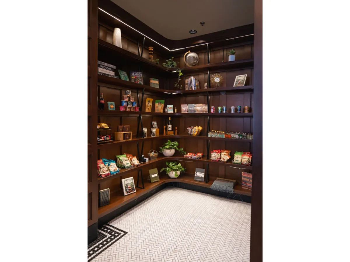 This cozy interior image features wooden shelves filled with locally sourced artisanal snacks, decorative plants, and textured floor tiles. The area invites guests to browse, relax with a coffee, and discover local products.