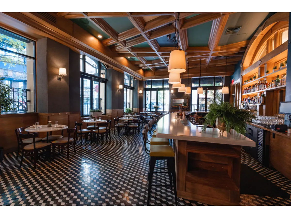 A lively, welcoming interior bar and dining area highlighted by a large bar counter, intricate ceiling design, checkerboard floor, and multiple dining tables. The atmosphere suggests a social, central hub where guests can gather, enjoy a drink, and relax after exploring the city with their pets.