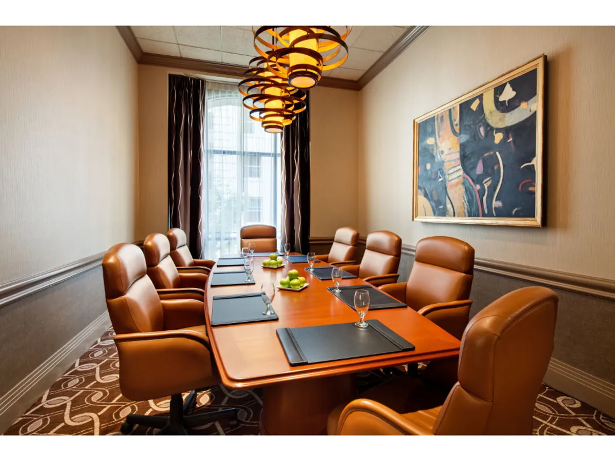 A spacious, modern conference room featuring a large wooden table, comfortable leather chairs, contemporary lighting, and art on the walls. Glassware on the table hints at meetings or brainstorming sessions, and the overall atmosphere is both formal and inviting.