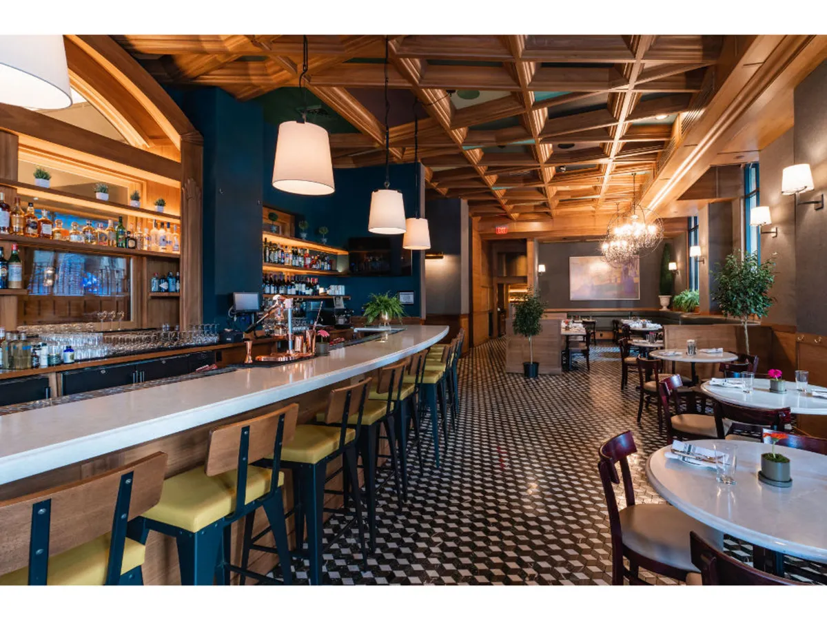 A stylish bar area featuring a long bar counter, unique hanging light fixtures, an ornate ceiling, and several dining tables. The atmosphere is both elegant and inviting, ideal for sipping handcrafted cocktails and enjoying small plates.