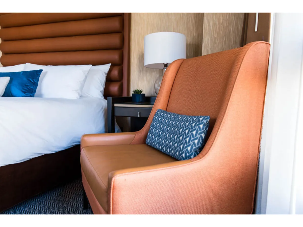 A cozy corner featuring an orange accent chair with a blue pillow, a plush bed dressed in white linens, and a modern lamp. The soft ambiance is enhanced by gentle natural light, making the setting perfect for relaxing, reading, or catching up on entertainment.