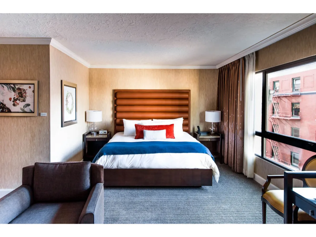 A chic bedroom with a plush king-sized bed adorned in blue and white bedding, accented with orange pillows. Beside the bed, modern lamps provide a warm glow, and stylish artwork brings a local touch. Natural light pours in through expansive windows, highlighting the cozy and contemporary decor.