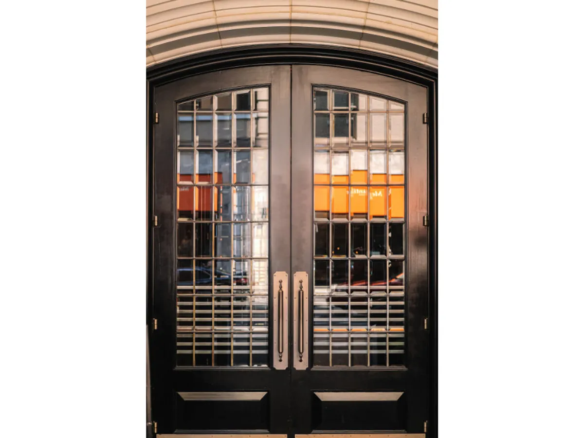 This exterior image showcases ornate double doors with grilled glass panels and metal handles, inviting guests into a bustling venue. There is a sense of movement and anticipation, as these doors mark the transition from the outside world into the vibrant atmosphere within.