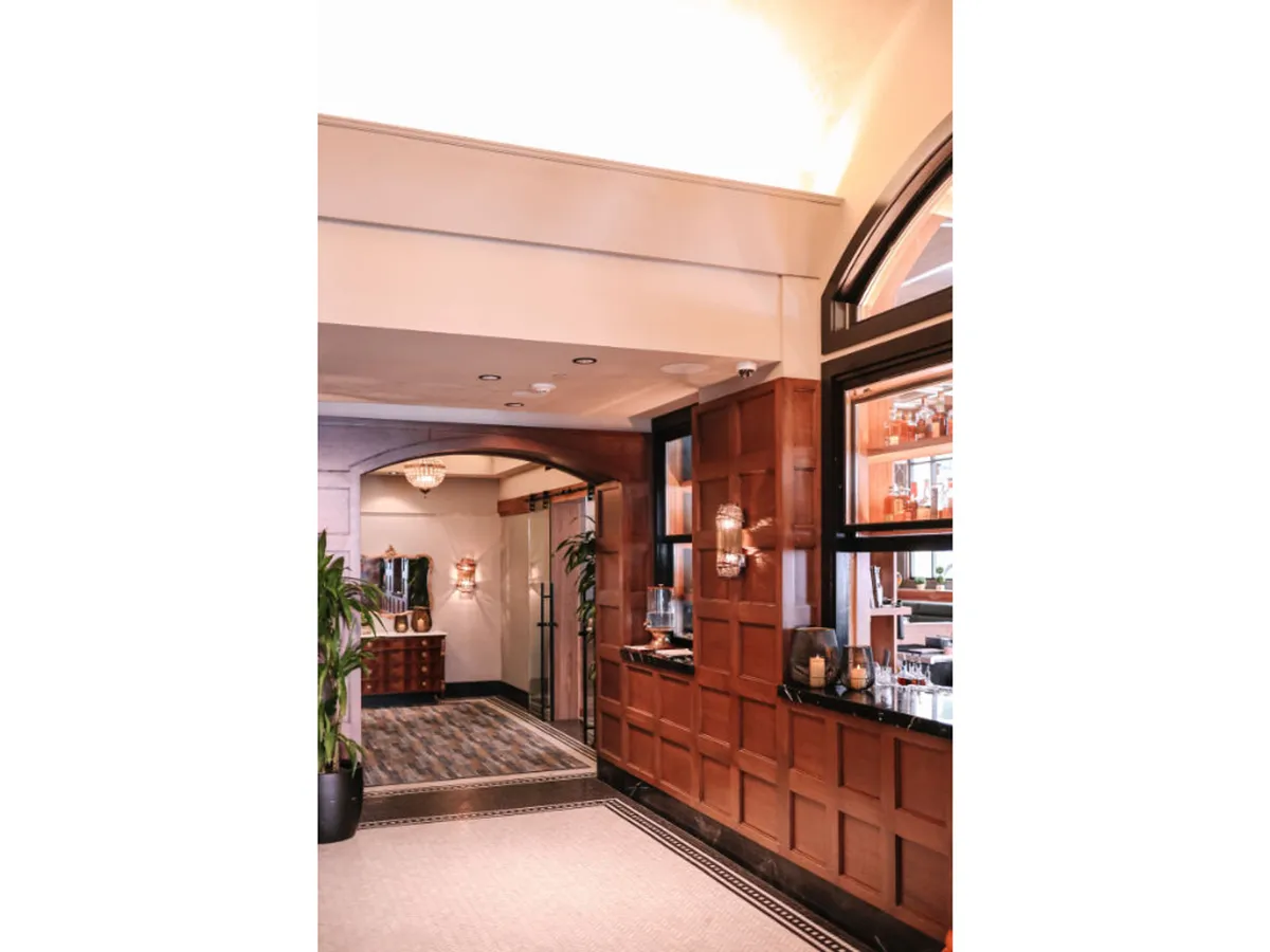 This image captures the sophisticated hotel lobby area, with wood paneling, elegant chandeliers, decorative lighting, a welcoming reception desk, and lush potted plants. There is a sense of arrival and luxury, with guests being greeted and others relaxing or socializing in the space.