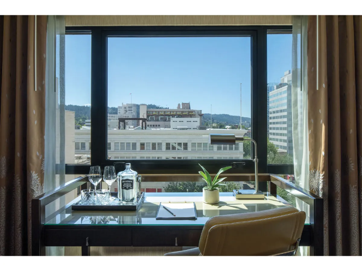 A modern workspace with a glass surface desk positioned by a large window, offering an abundance of natural light. On the desk are a bottle and two glasses, along with a vibrant green plant, creating a relaxed yet productive setting. The scene suggests both work and leisure possibilities, with a view that inspires focus or a calming break.