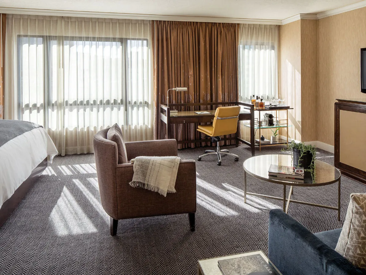 A modern bedroom featuring a plush bed with crisp white linens, a sleek desk with a yellow chair, and large windows draped with sheer curtains. A small coffee table displays decorative items, and the space feels both relaxing and functional, ideal for enjoying city views or a morning coffee.